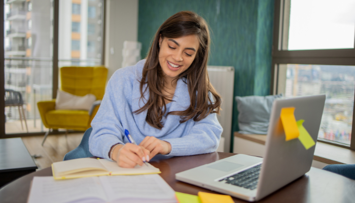 Young woman studying at a desk with a laptop, notebook, and sticky notes in a bright, modern workspace. She is smiling while writing, showcasing a productive study environment.